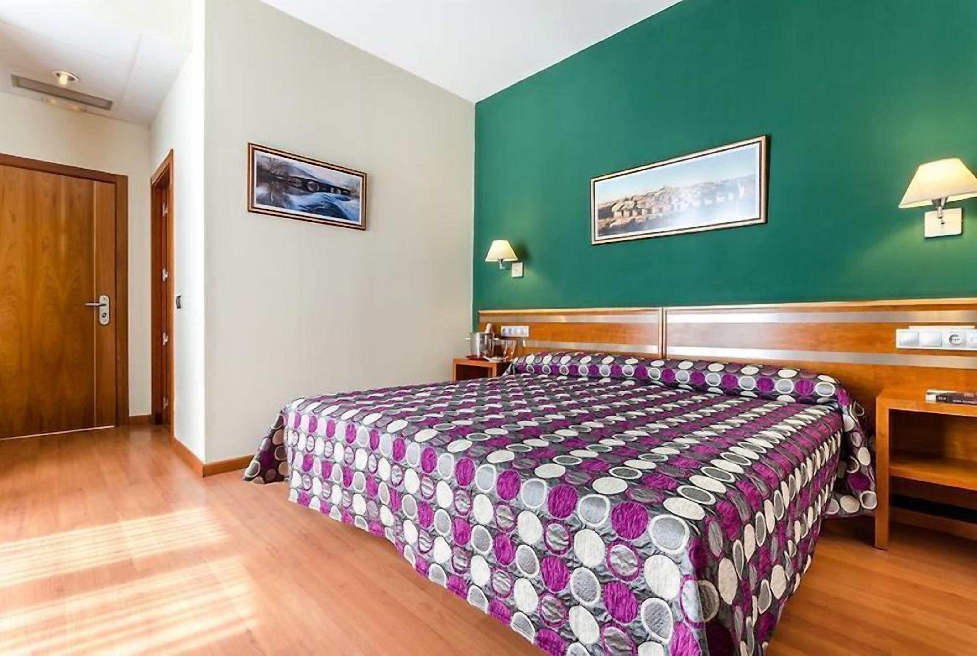 Una habitación de hotel con una cama grande, un cabecero de madera, dos mesitas de noche, una pared de acento verde y dos cuadros enmarcados.
