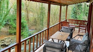 Een overdekt terras met houten leuningen, een stenen muur en uitzicht op een bebost gebied.