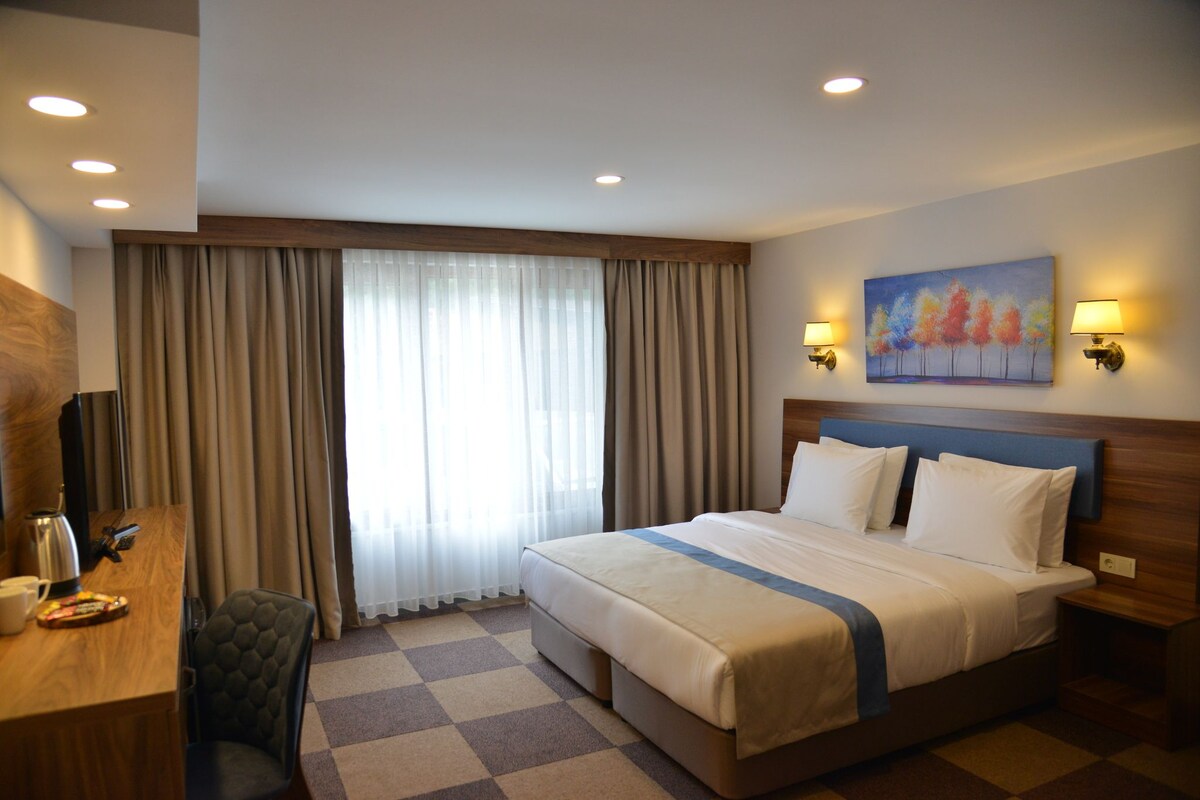 A hotel room with a large bed, a desk with a chair, a painting on the wall, and a window with curtains.