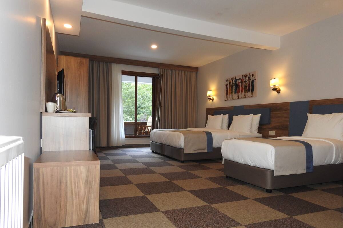 Een hotelkamer met twee bedden, een zwart-wit geruite vloer, een koffiezetapparaat en uitzicht op groen door het raam.