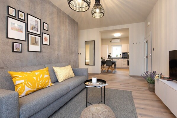 A modern living room with a grey sofa, yellow and white cushions, a small round table with a magazine, and a grey rug. There are framed pictures on the wall and a kitchen area in the background.