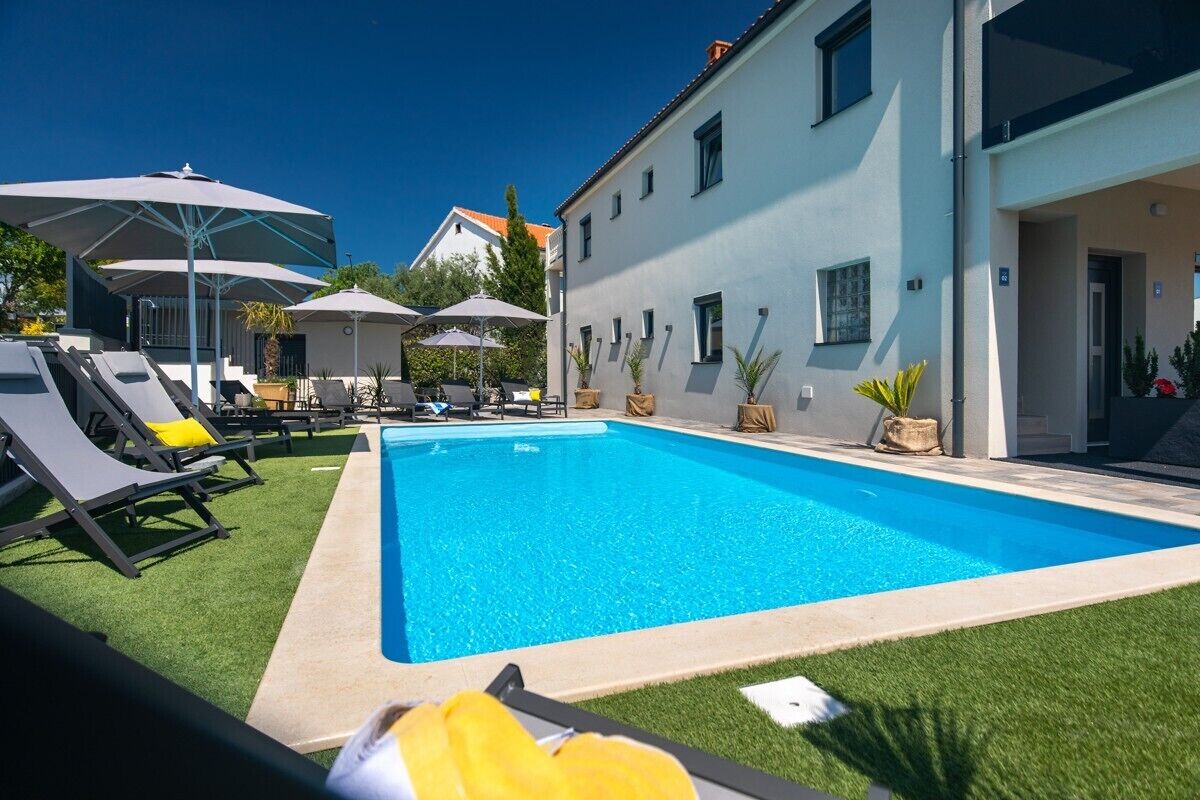 Una piscina con sillones de descanso y sombrillas, rodeada por un edificio de fachada azul claro.
