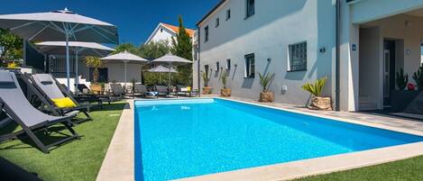 Una piscina con sillones de descanso y sombrillas, rodeada por un edificio de fachada azul claro.