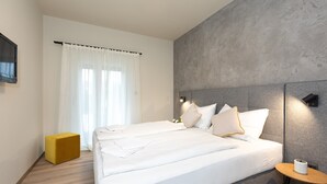 Ett modernt hotellrum med en stor säng, en liten gul pall, ett nattduksbord med en lampa och ett stort fönster med tunna, genomskinliga gardiner.