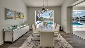 A modern dining area with a glass-top table, white chairs, and a view of a pool through a large window. - Villa Inspiration - modern style, spectacular view, large pool area  (Cape Coral)
