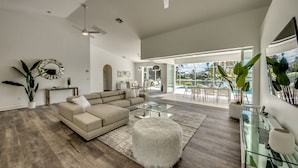 A spacious living room with a large sectional sofa, a glass coffee table, and a white fluffy ottoman. There is a dining area with a glass table and chairs visible through the open space. - Villa Inspiration - modern style, spectacular view, large pool area  (Cape Coral)