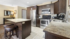 A modern kitchen with dark wood cabinets, stainless steel appliances, a granite countertop island, and a bar stool. - Townhouse with Free Shuttle to Tremblant Resort (Mont-Tremblant)