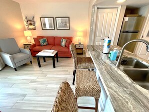 Un salon comprenant un canapé, des fauteuils, une table basse et un coin cuisine avec un évier et un plan de travail.