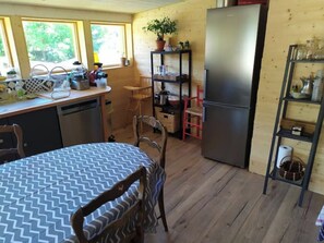 A compact kitchen with a dining area, wooden walls, a refrigerator, and a shelf with various items.