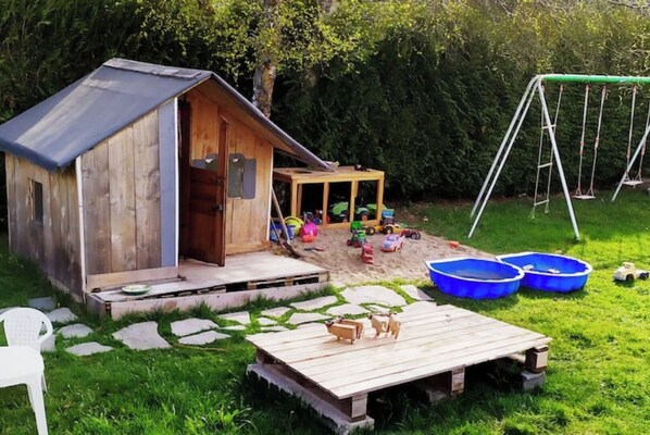 A wooden shed with a door, a small play area with toys and a swing set, and a wooden deck with two blue pools.