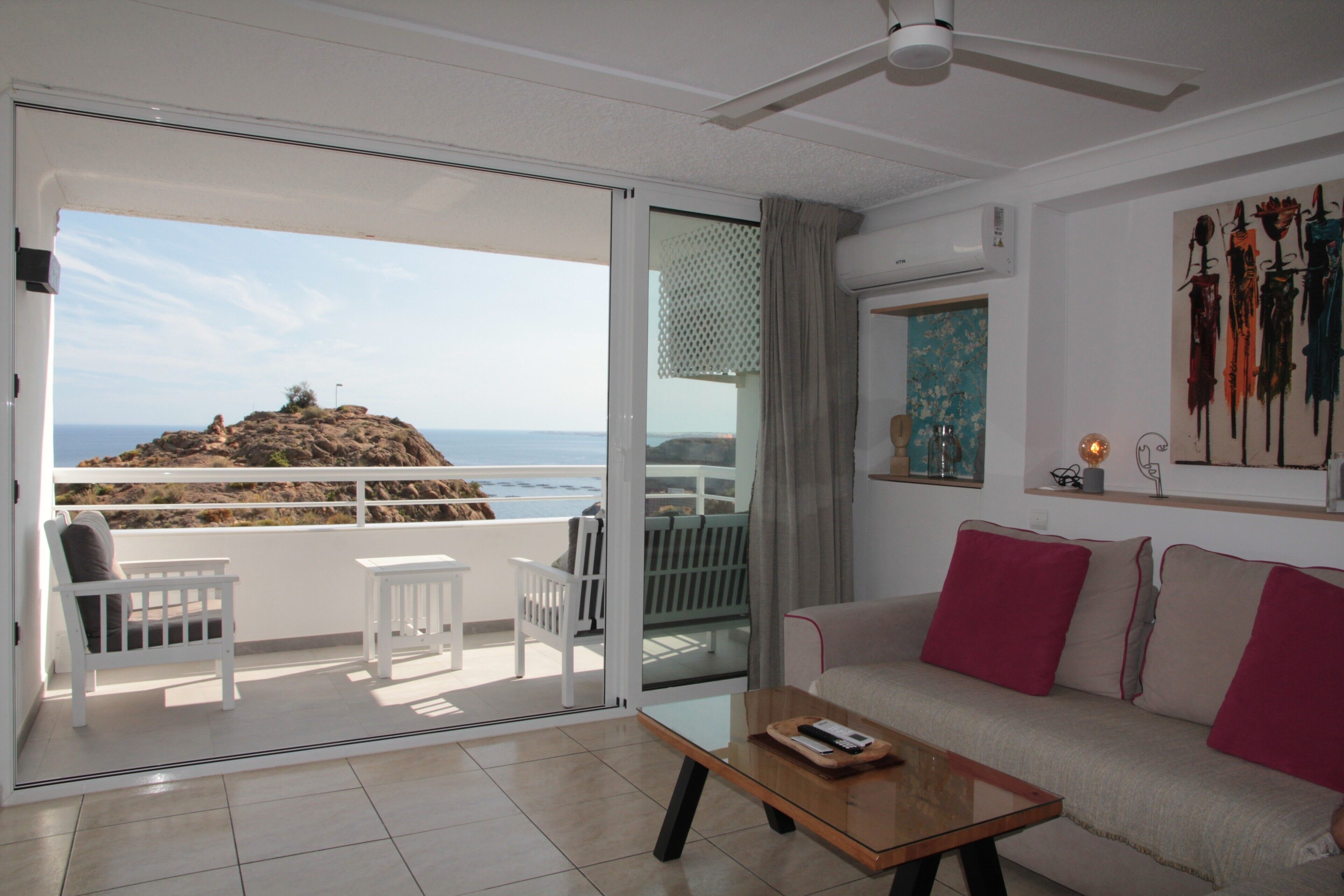 Una stanza con divano, tavolino, balcone con sedie e vista sul mare.