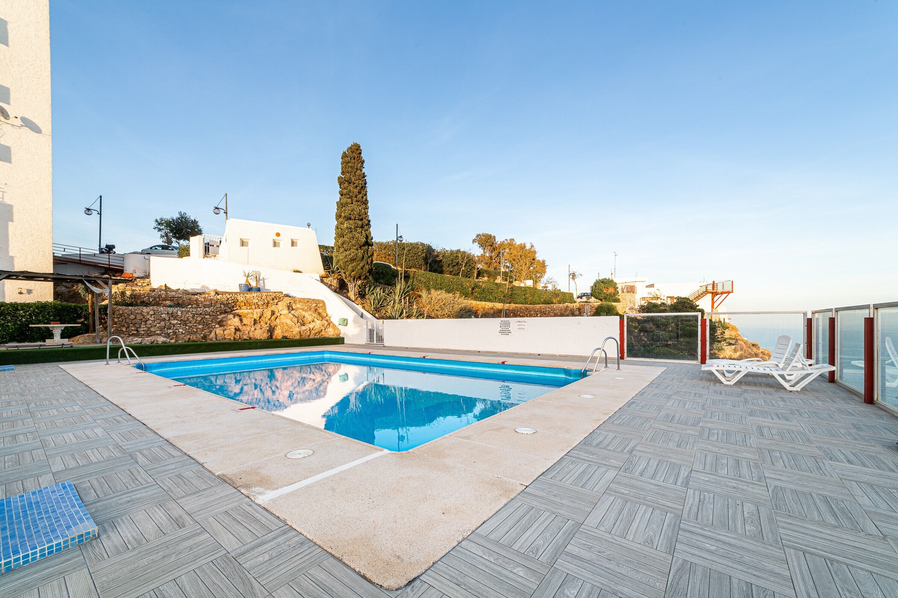 Une piscine avec une terrasse au motif en chevrons, un espace détente avec des chaises blanches et une vue sur un paysage côtier.