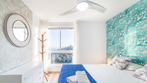 Une chambre à coucher avec un lit, une couvre-lit bleue, un mur blanc orné d’un motif floral, un miroir rond et une fenêtre donnant sur un paysage.