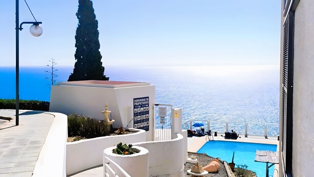 Un espace piscine offrant une vue dégagée sur la mer, un panneau indiquant le nom de la communauté de propriétaires, et un grand arbre.
