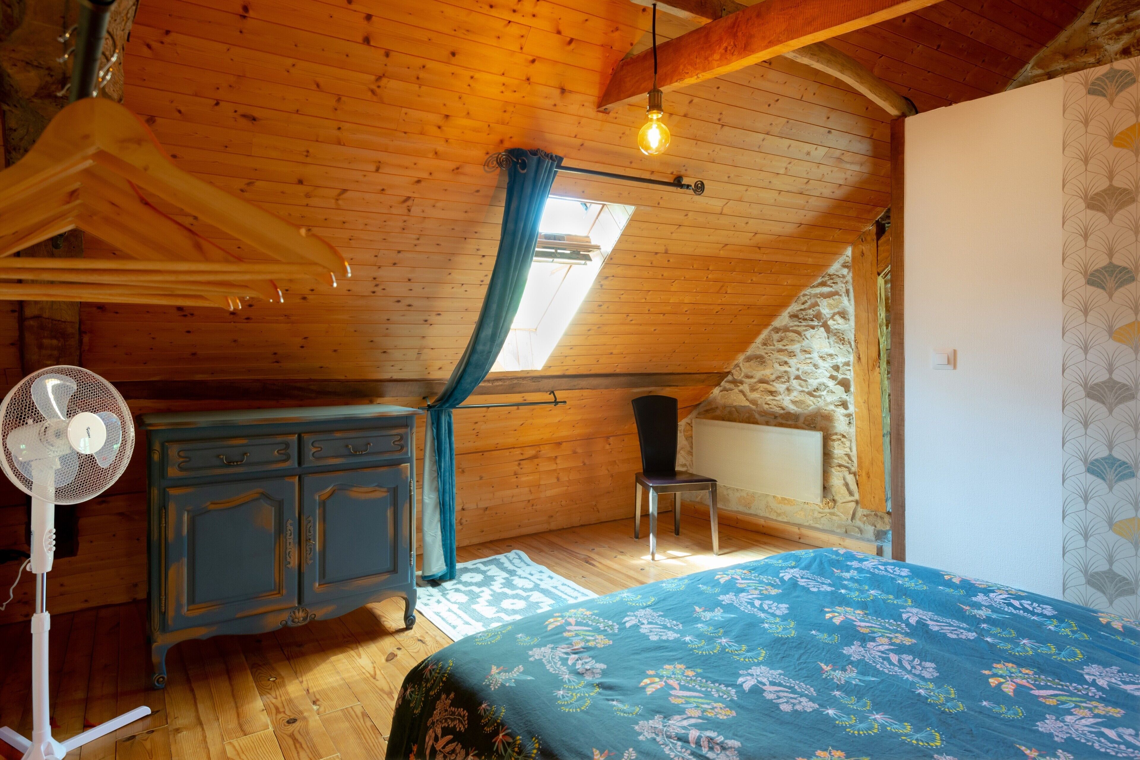 A room with a sloped ceiling, a bed with a floral bedspread, a dresser, a chair, a fan, and a window with a curtain.
