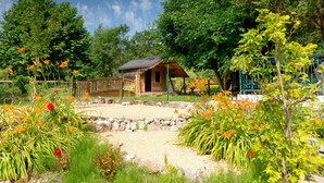 Un chalet rustique doté d’une terrasse en bois, entouré d’un jardin où poussent différentes plantes et fleurs.