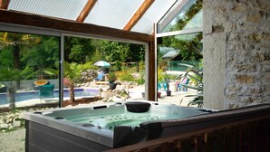 Un bain à remous avec une terrasse en bois à l’avant, un mur de pierre sur la droite et une vue sur une piscine et un jardin, visibles à travers de grandes fenêtres.