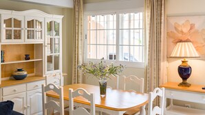 A dining area with a wooden table, white chairs, a vase with flowers, and a window with curtains. - Holiday Home La Reserva de Alvarito with Pool, Garden, Terrace & Wi-Fi (Marbella)