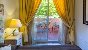 A room with a bed, a bedside lamp, a window with yellow curtains, and a view of an outdoor patio with chairs and a table. - Holiday Home La Reserva de Alvarito with Pool, Garden, Terrace & Wi-Fi (Marbella)