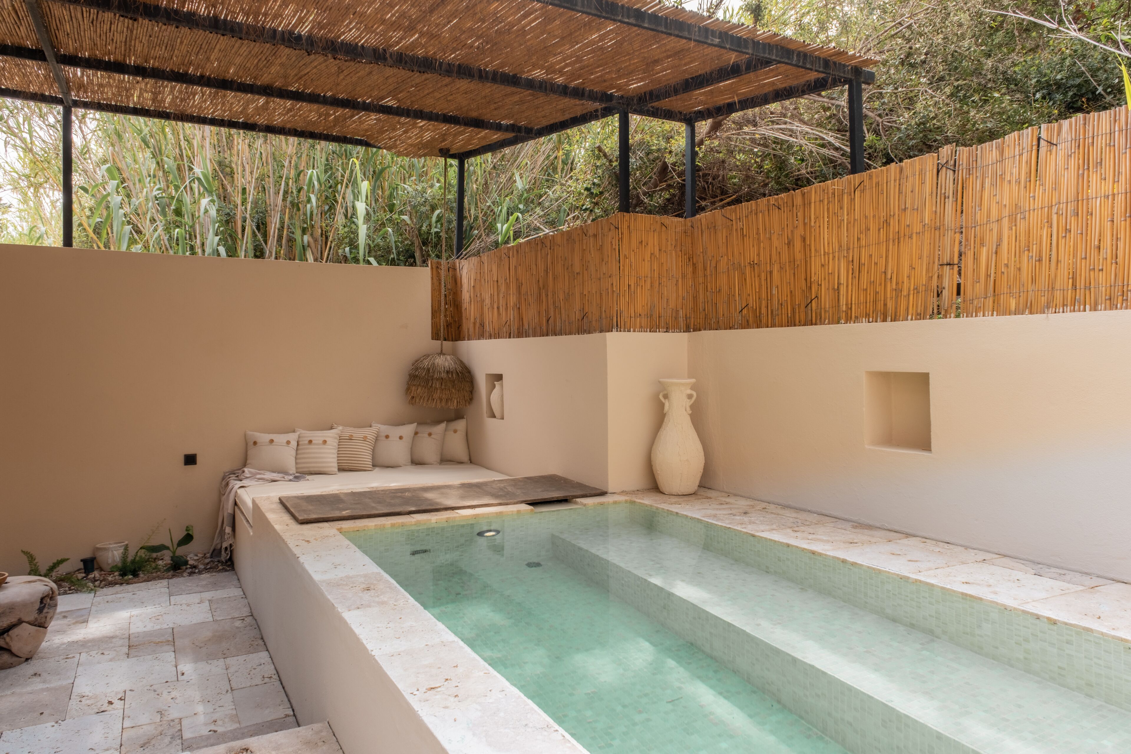Un área de piscina moderna al aire libre con una pared de bambú, una zona de estar con cojines y un jarrón grande.