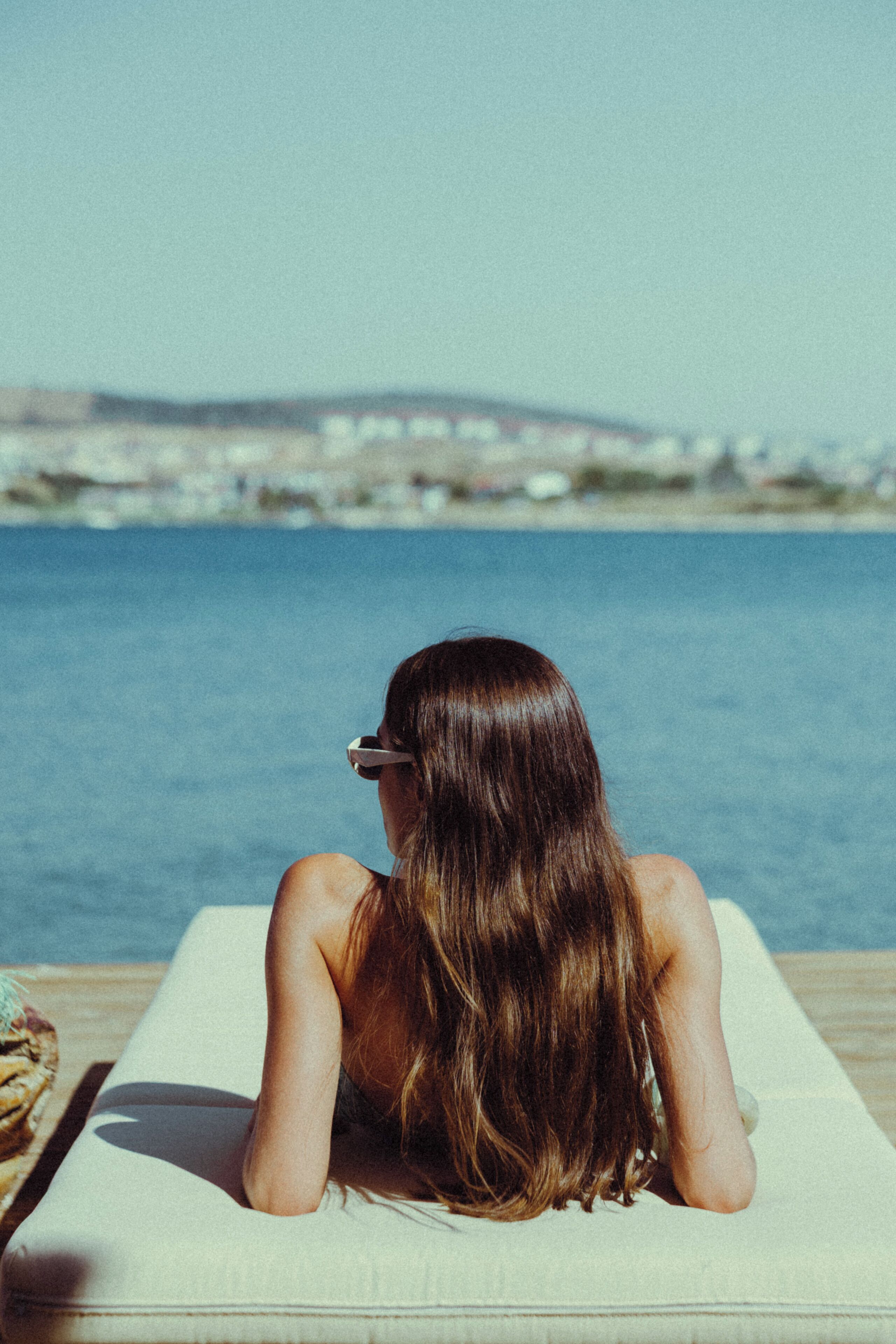 A person reclining on a sun lounger by a body of water with a distant shoreline.