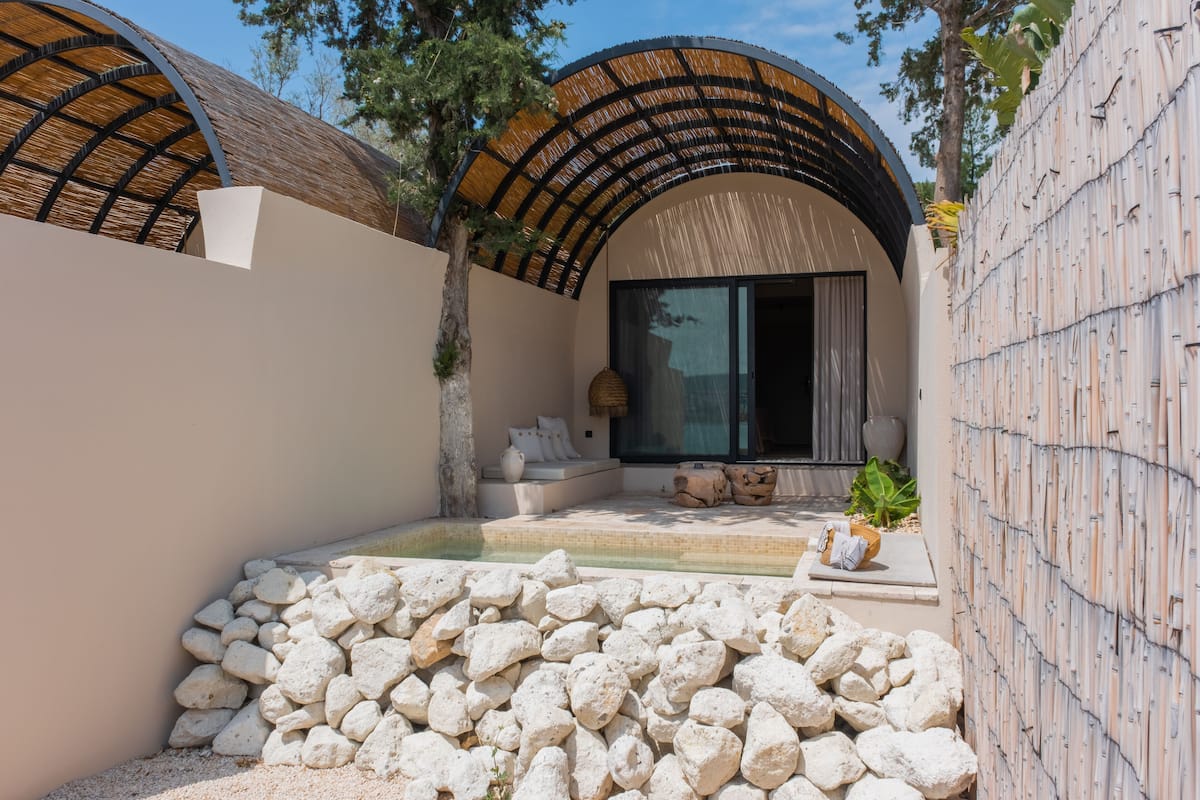 A modern outdoor patio with a stone wall, a seating area with cushions, and a large window.