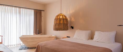 A modern hotel room with a large bed, a wooden side table, a hanging light fixture, and a large window with curtains.