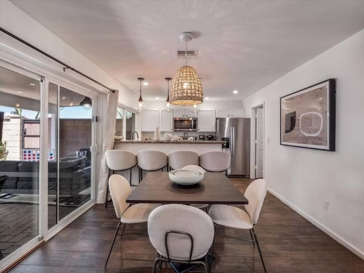 A modern dining area with a large table, multiple chairs, a chandelier, and a kitchen in the background.