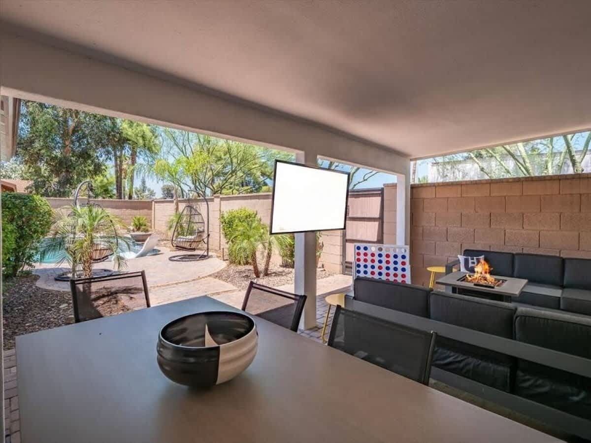 Um pátio ao ar livre com mesa, cadeiras, lareira e vista para uma piscina e vegetação.