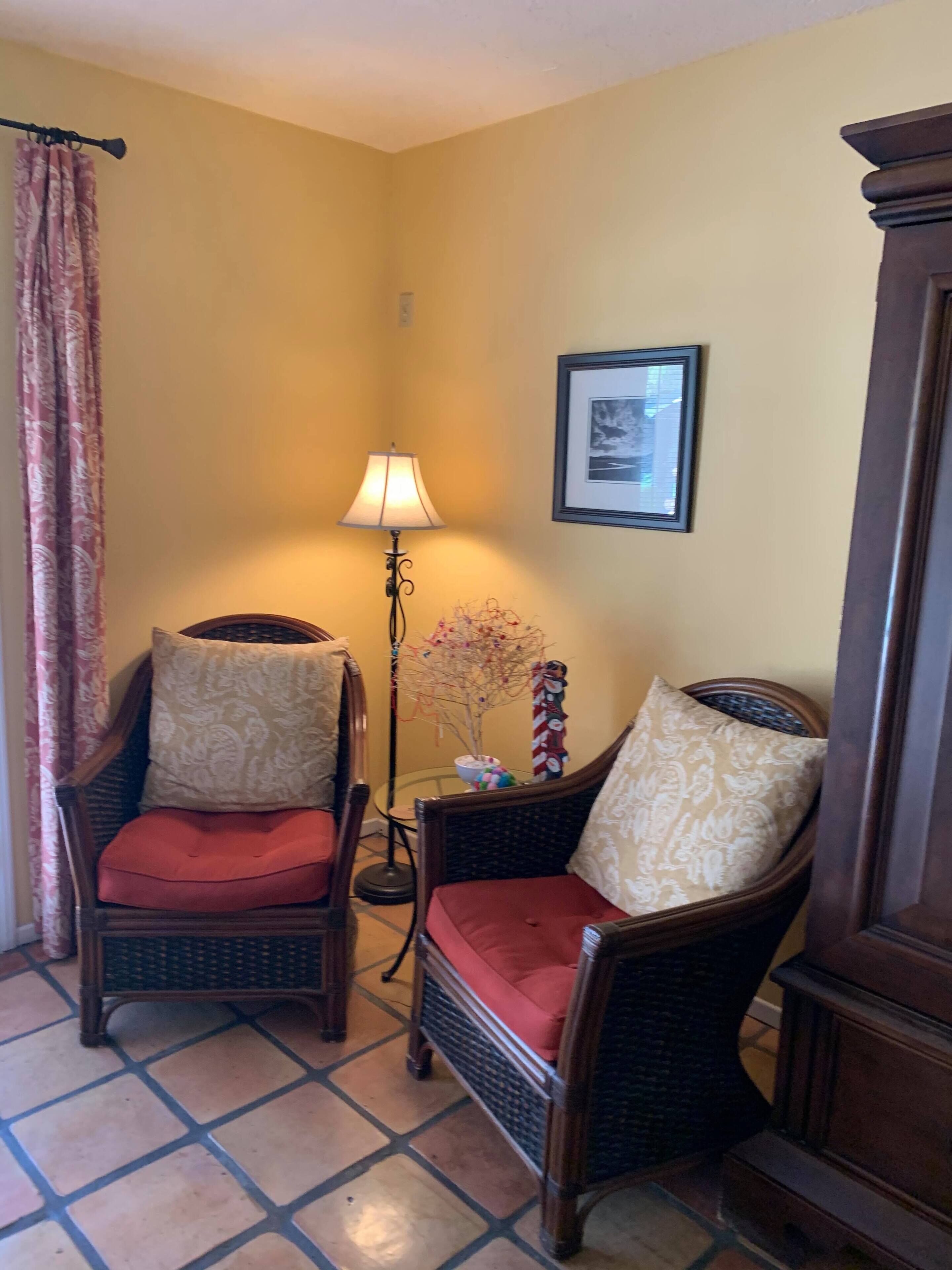 Una habitación de esquina con dos sillones de mimbre y cojines estampados, una lámpara de pie, un cuadro en la pared y una cortina.