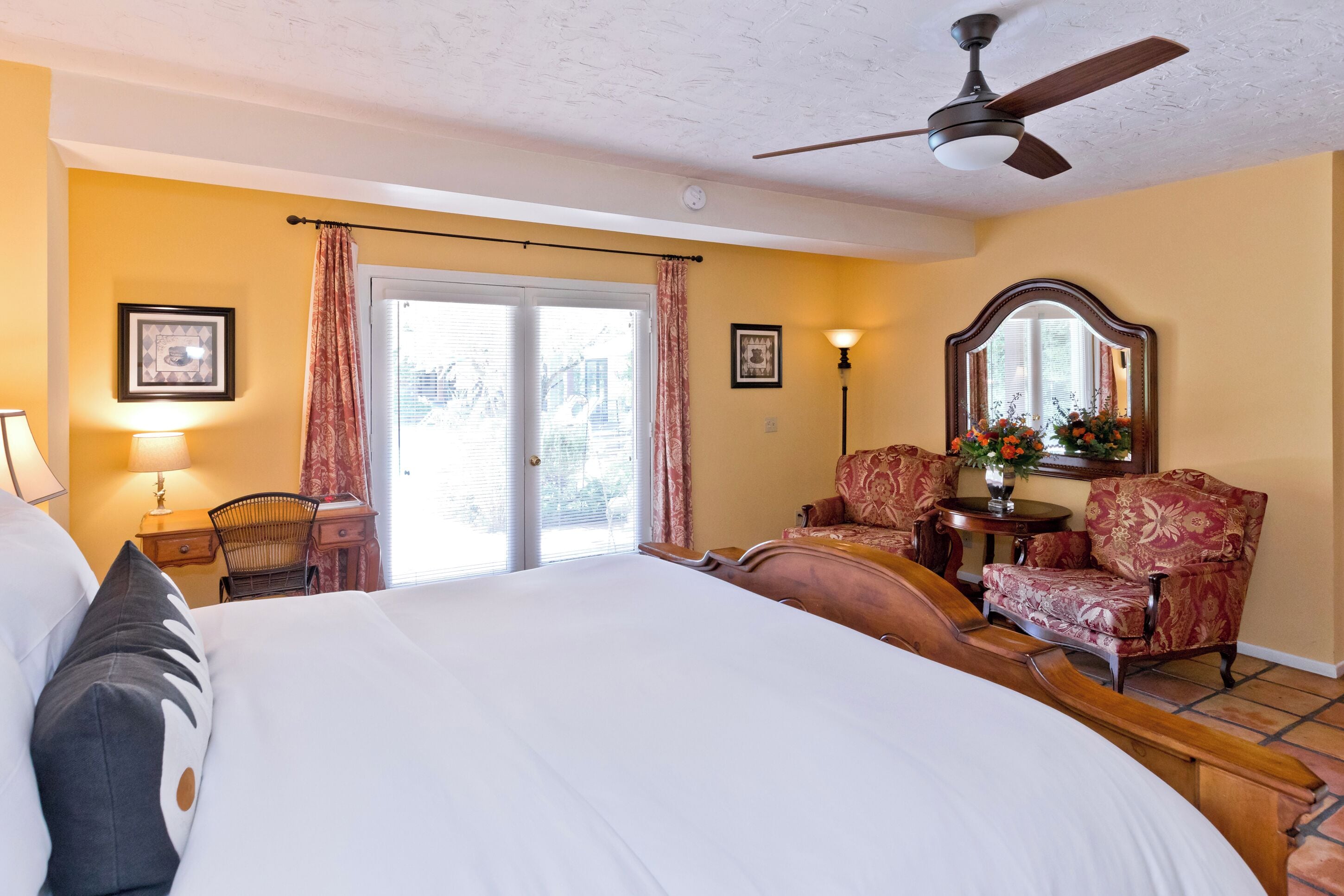 Une chambre d’hôtel avec un grand lit, un bureau avec une chaise, une lampe de bureau, un miroir et une fenêtre avec des rideaux.
