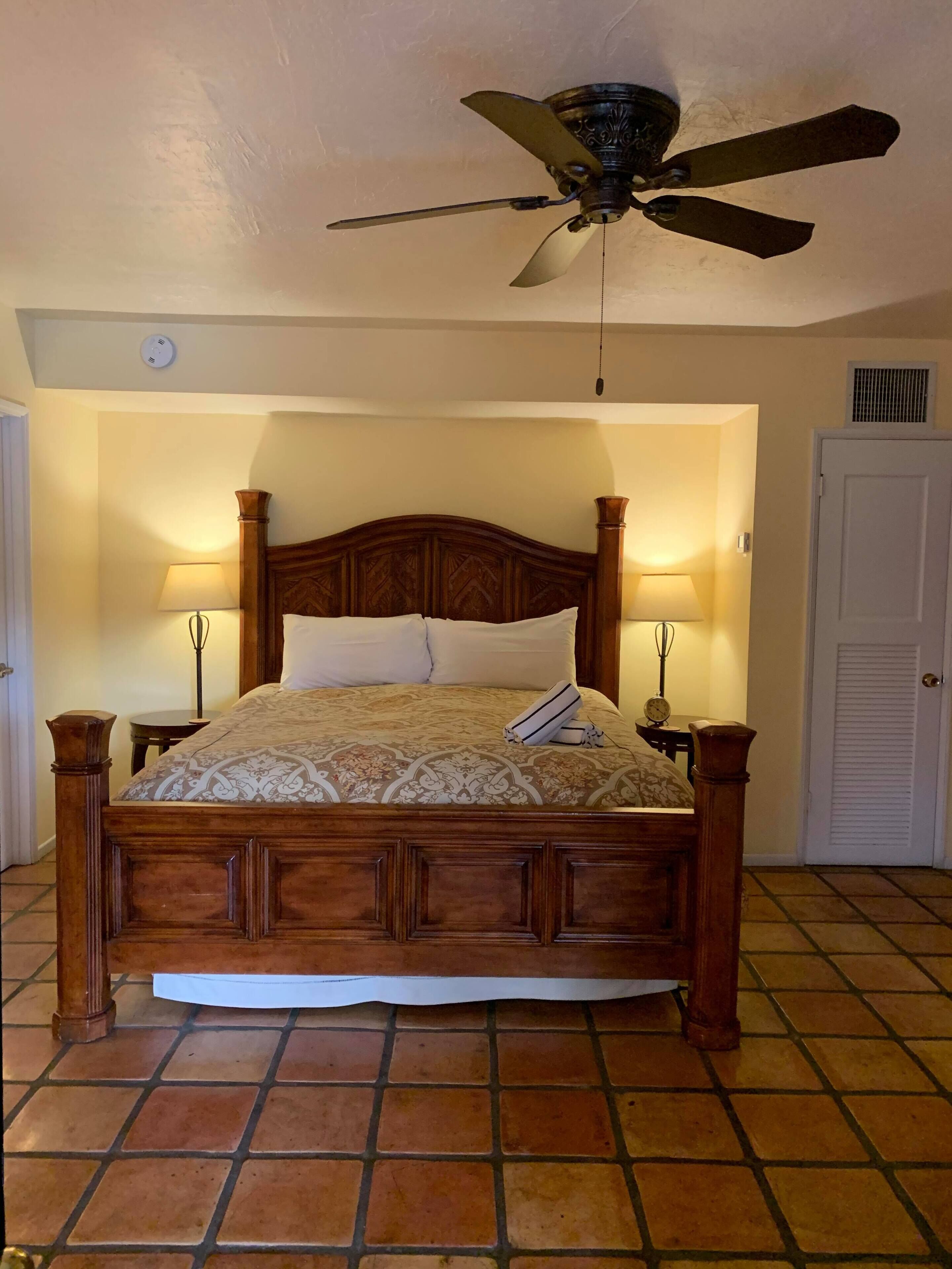 A bedroom with a wooden bed, two bedside lamps, a ceiling fan, and a tiled floor.