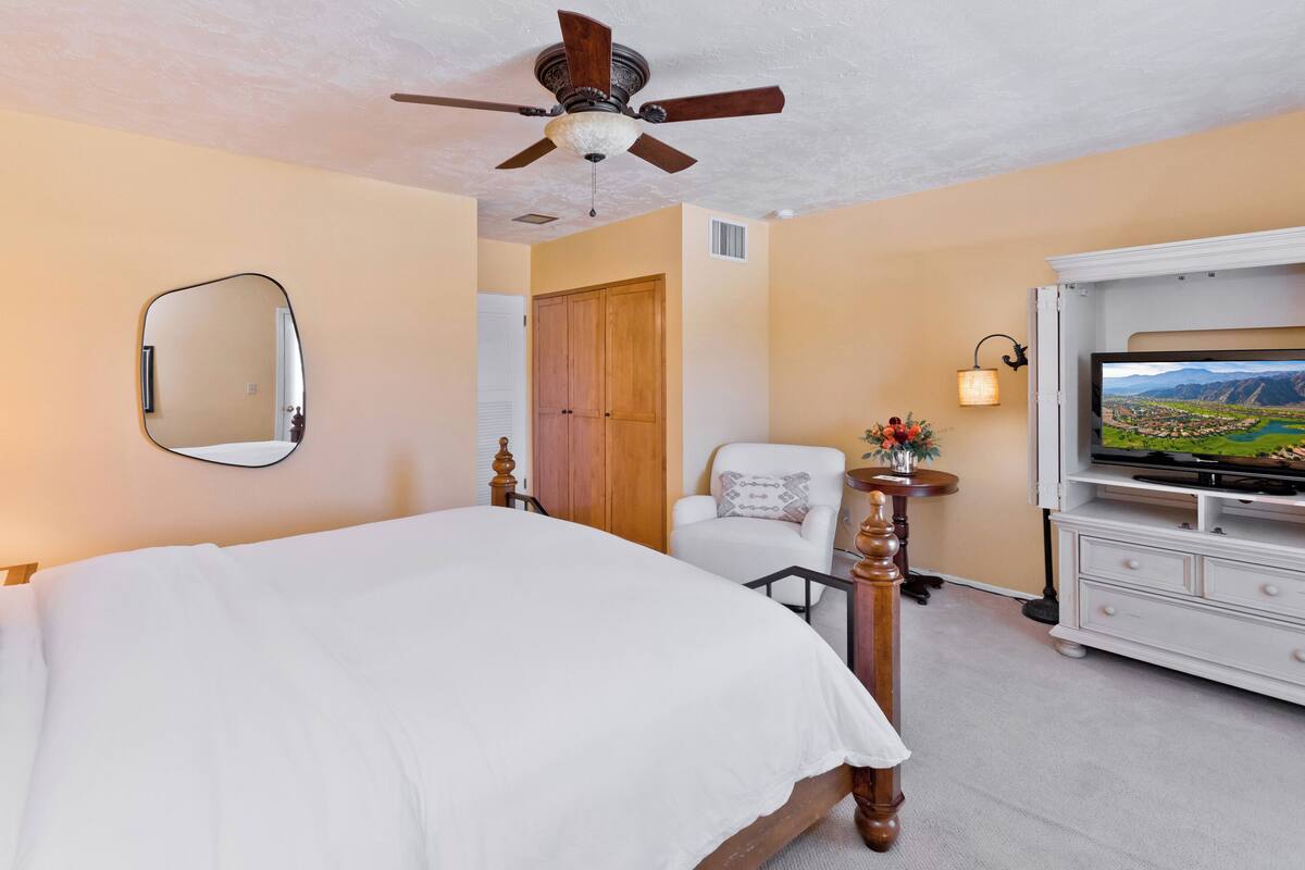 A hotel room with a bed, a ceiling fan, a television, a chair, a dresser, and a mirror.