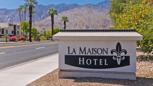A sign for La Maison Hotel with a fleur-de-lis logo, set against a backdrop of mountains and palm trees. - LaMaison Palm Springs (Palm Springs)