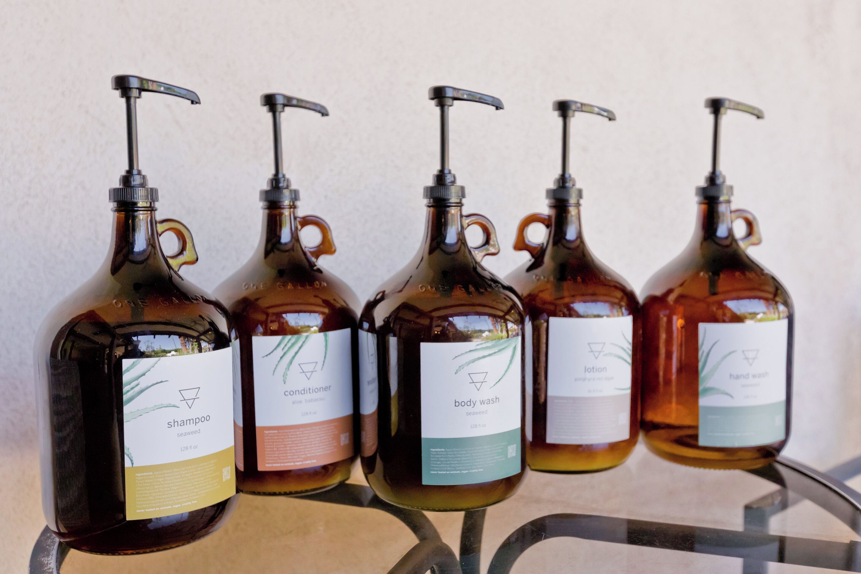 Four glass bottles with pumps, each labeled with different product names like shampoo, conditioner, body wash, and hand wash.