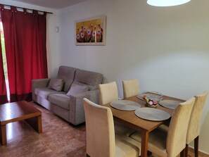 A living room with a sofa, a dining table with chairs, a painting on the wall, and a red curtain.