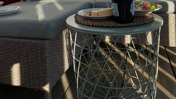 A wicker sofa with cushions, a glass table with a bottle and glasses, and a plate of food.
