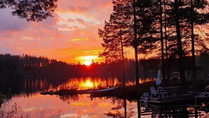 A sunset over a calm lake with a dock and chairs, surrounded by trees.