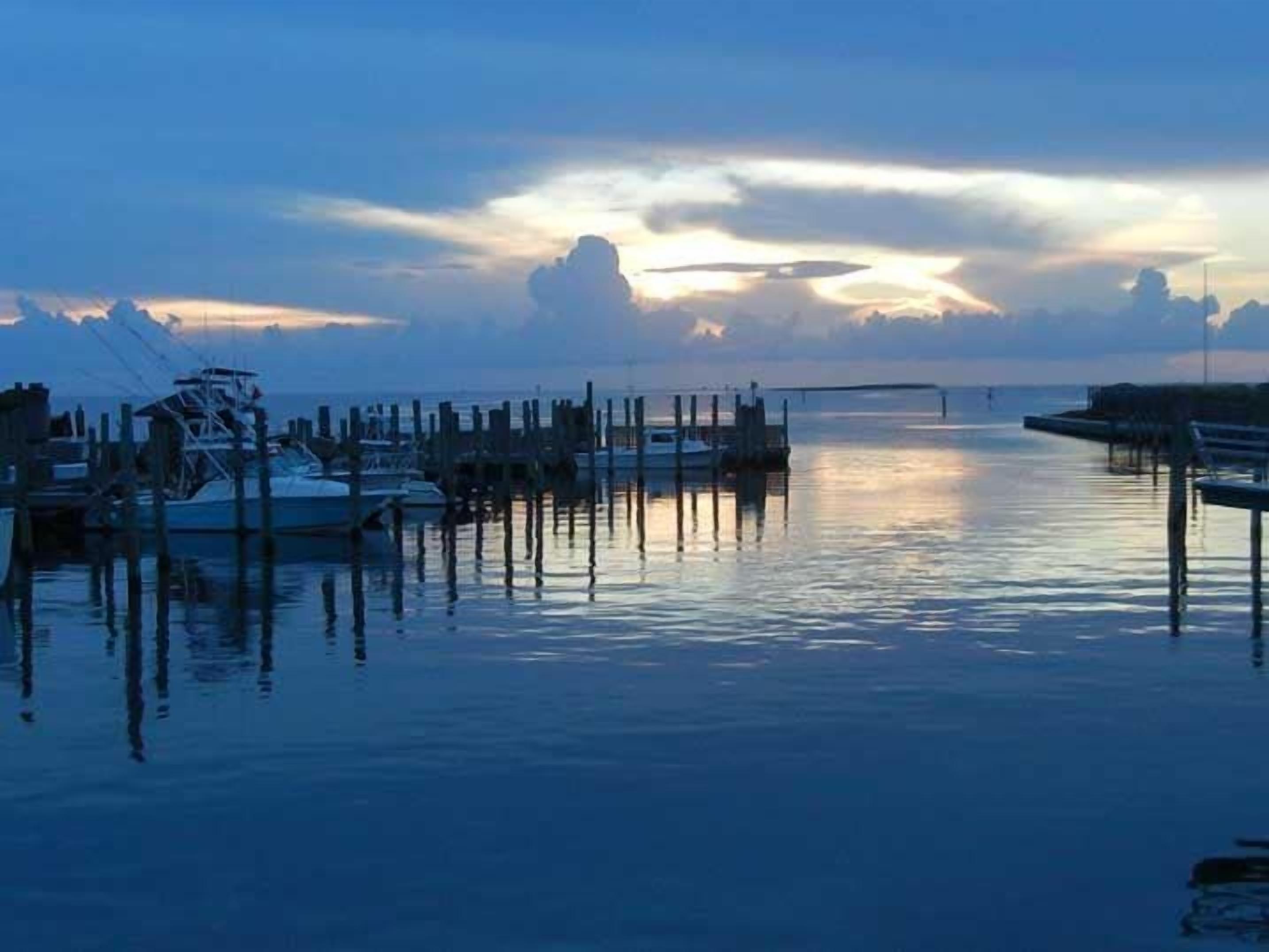 Un puerto deportivo con barcos amarrados a muelles de madera, agua tranquila y un atardecer de fondo.