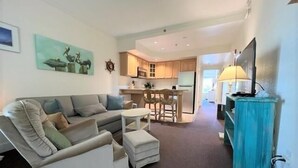 A living room with a sofa, ottoman, and a TV stand. There is a kitchen in the background with a dining area. - Just Beachy!  Short walk to the beach, shops and ferry! (Hatteras)