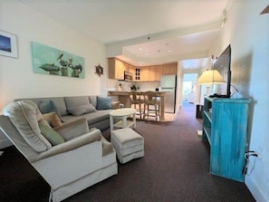 A living room with a sofa, ottoman, and a television on a stand. - Charming one bedroom condo with private patio. HI-101 (Hatteras)