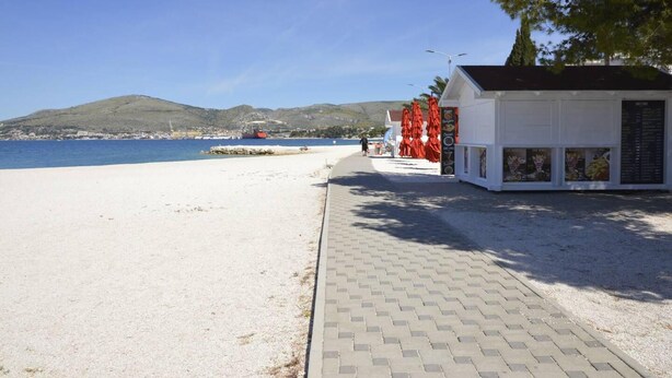 A beachside walkway leading to a white building with red umbrellas, a clear blue sky, and a distant view of a red boat on the water. - Stunning 2-bed Apartment in Okrug Gornji (Okrug)
