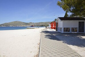 A beachside walkway leading to a white building with red umbrellas, a clear blue sky, and a distant view of a red boat on the water. - Stunning 2-bed Apartment in Okrug Gornji (Okrug)