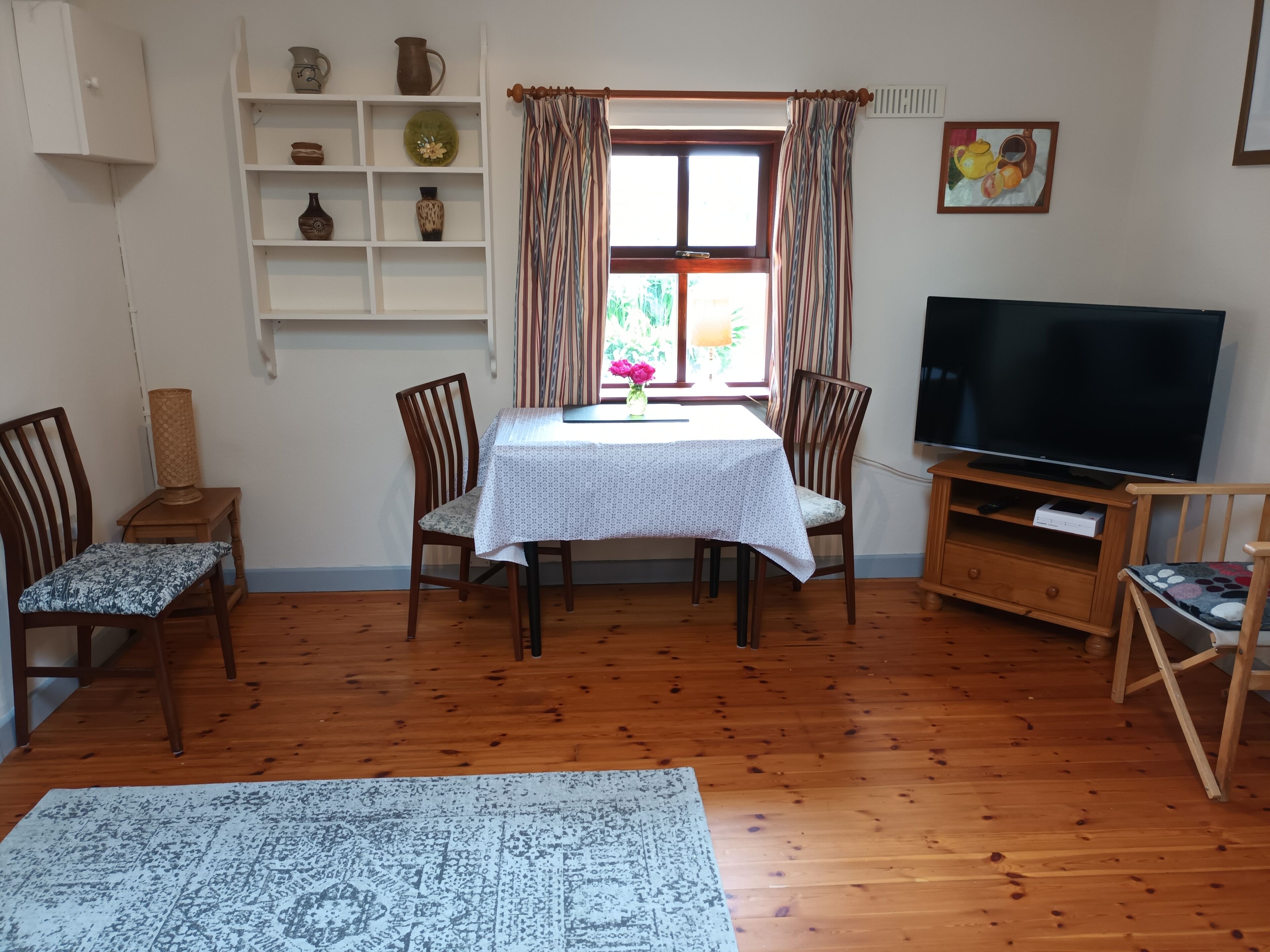 A living room with a wooden floor, a dining table with chairs, a TV on a wooden stand, and a shelf with decorative items.