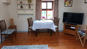 A living room with a wooden floor, a dining table with chairs, a TV on a wooden stand, and a shelf with decorative items.