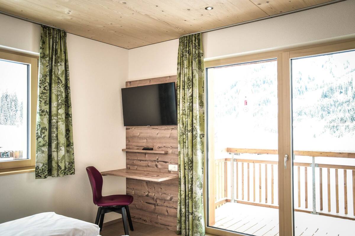 A room with a wooden ceiling, a TV mounted on a wooden shelf, a red chair, and a balcony with a wooden railing.