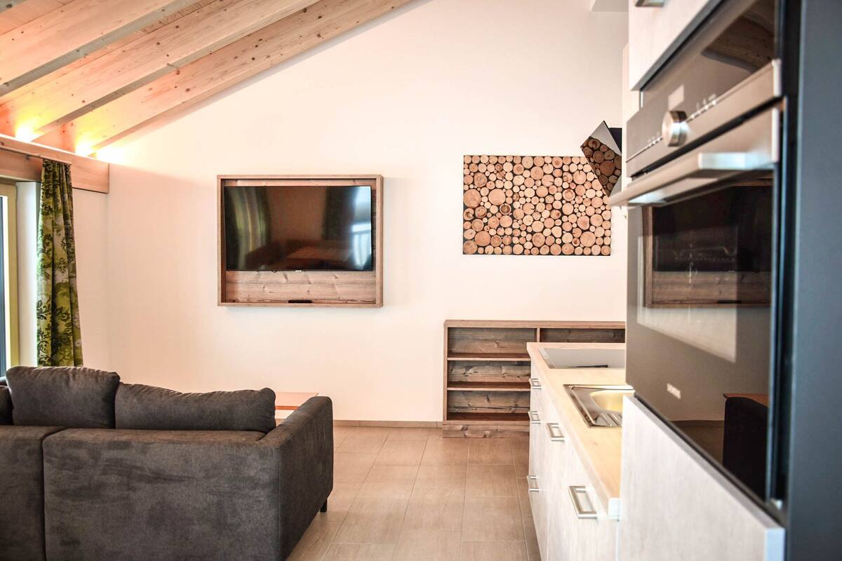 A modern kitchen with a built-in oven, a wooden wall art piece, and a grey sofa.