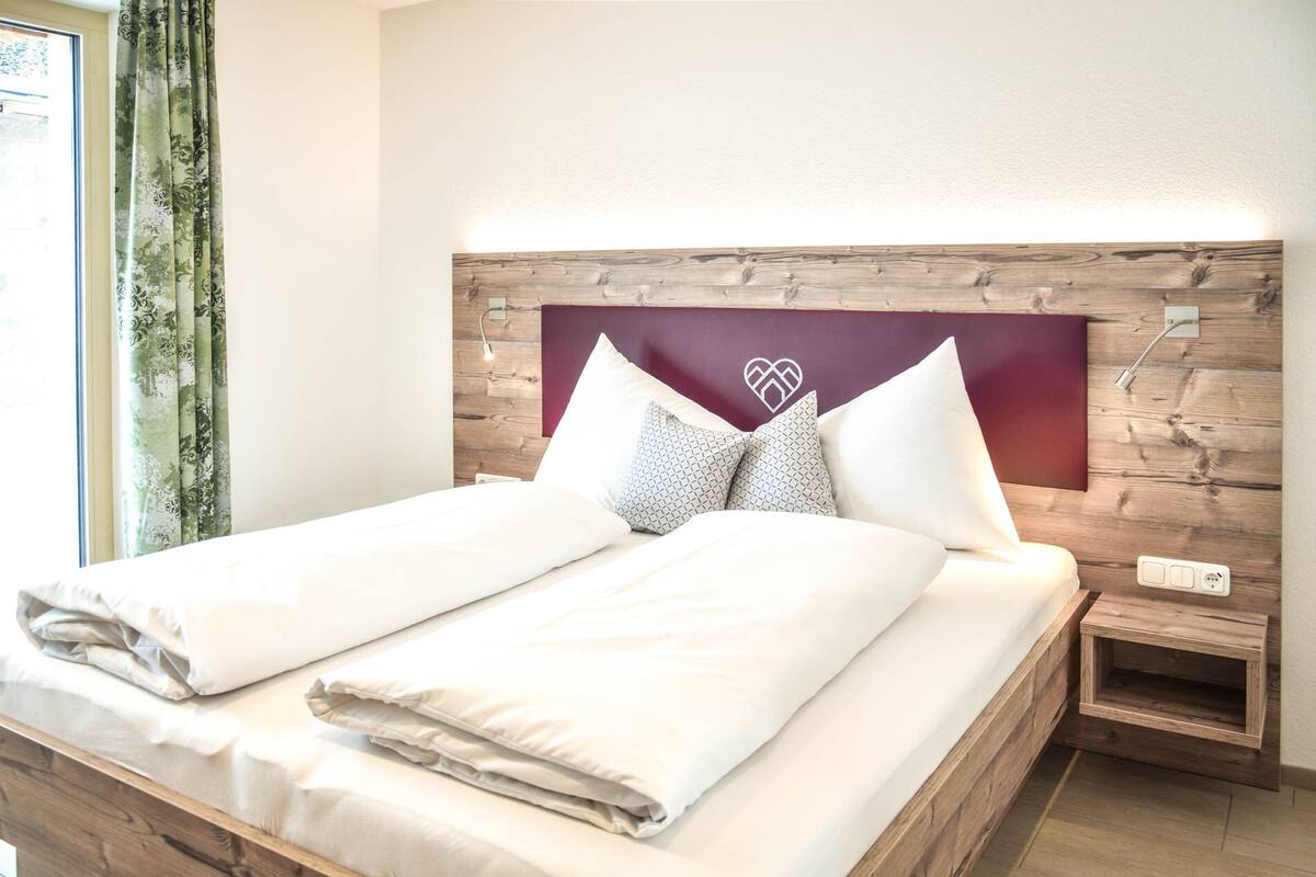 A neatly made bed with white linens and patterned pillows, a wooden headboard with a heart design, and a small bedside table with a lamp.