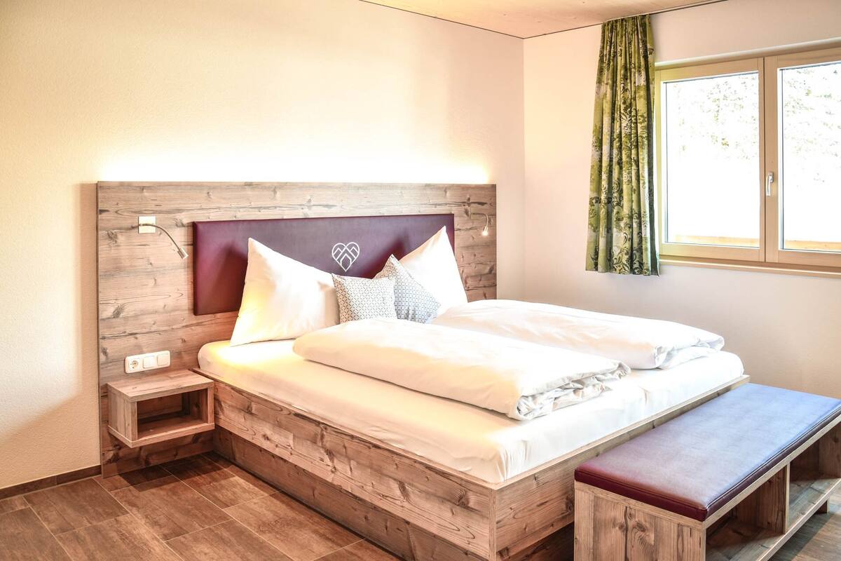 A bedroom with a wooden headboard, a bed with white bedding, a bedside table, a bench, and a window with curtains.