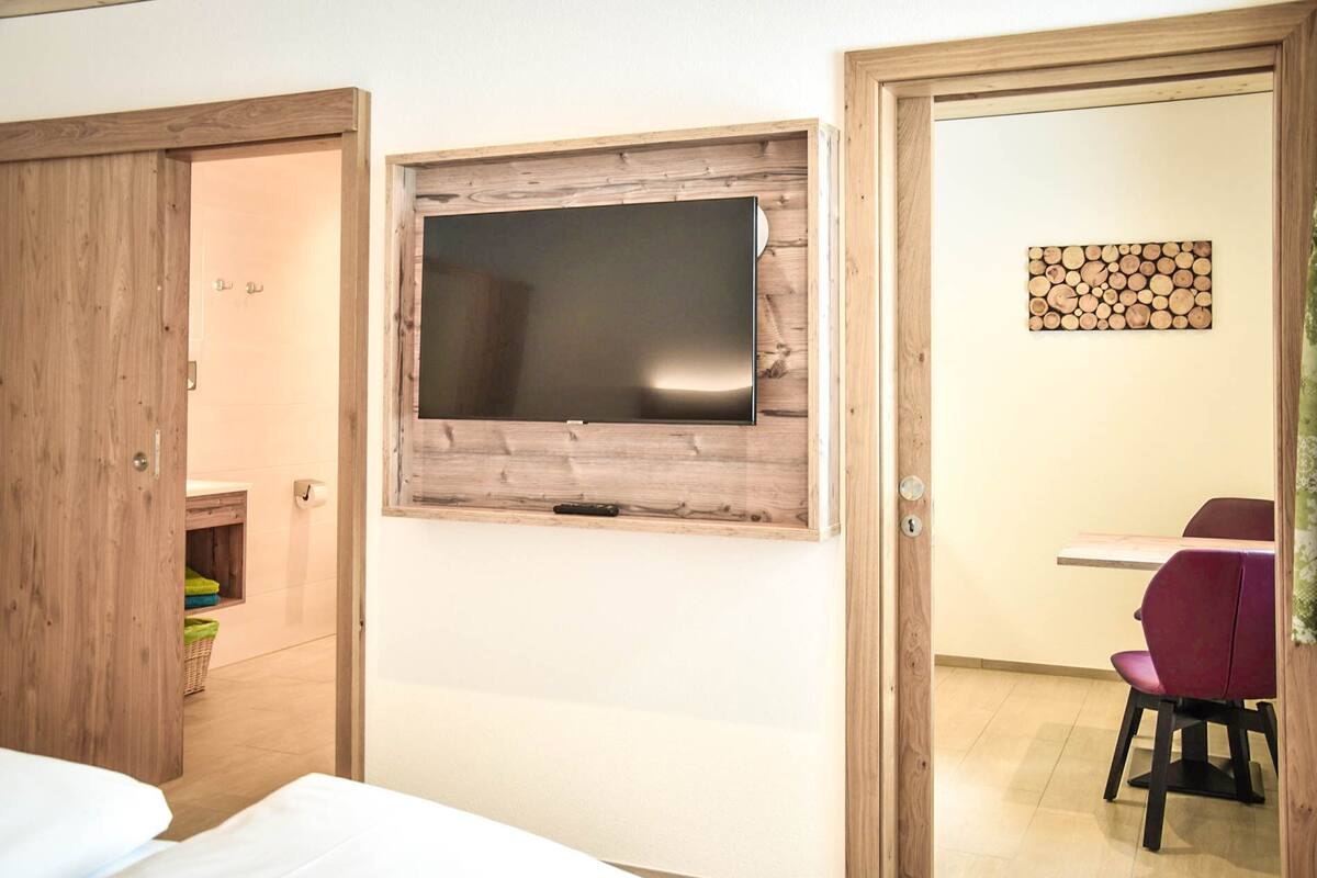 A room with a wooden-framed TV mounted on the wall, a wooden door, a small table with a chair, and a wall-mounted log art piece.